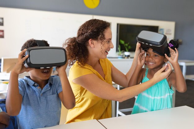 Caucasian young female teacher wearing vr glasses to african american elementary schoolgirl in class