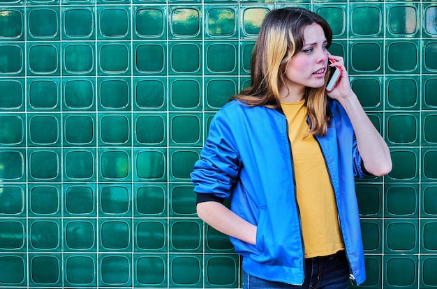 Caucasian young girl talking on the mobile phone over a green wall in the street