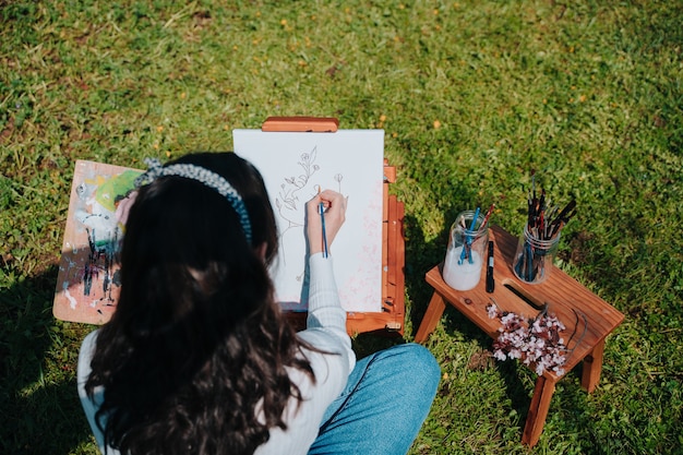 巨大なピンクのアーモンドの木の前で、公園の自然の中に立っている間、パステルピンクの絵の具でキャンバスを描いているウェーブのかかった髪の白人の少女。