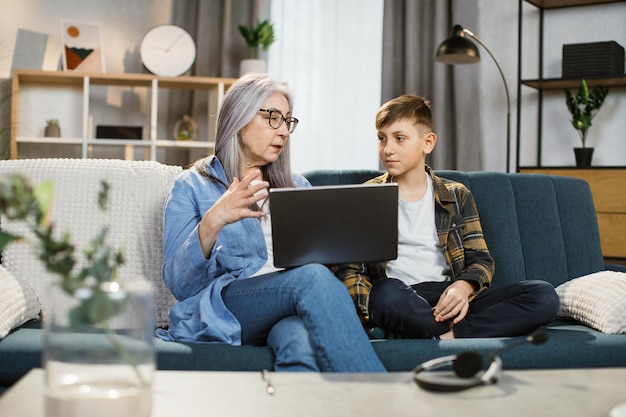 Caucasian young handsome boy teaching his old grayhaired grandmother to use laptop on couch