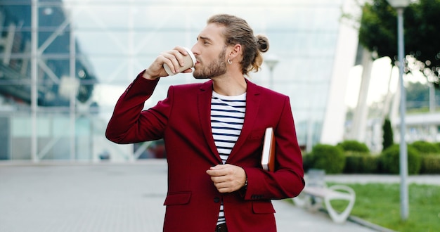 Caucasian young handsome stylish businessman walking outdoor in city with planner and sipping drink. Business center modern building on background. Male strolling and drinking coffee to-go.