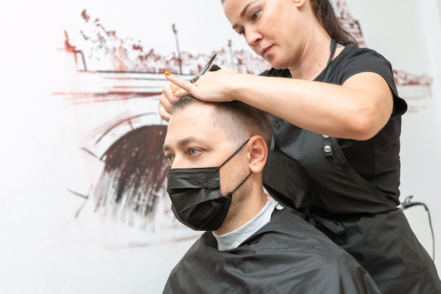 保護マスクを身に着けている理髪店で髪を切っている白人の若い男。ニューノーマル。社会的距離。