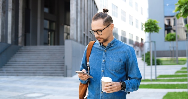 Caucasian young man in glasses tapping and scrolling on mobile phone and holding coffee to-go at city street. Handsome male in eyeglasses texting message on smartphone outdoors. Gadget using concept.