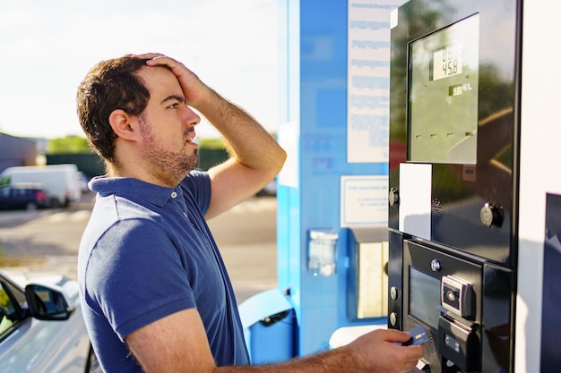 Caucasian young man surprised by high fuel prices on the gas station scoreboard