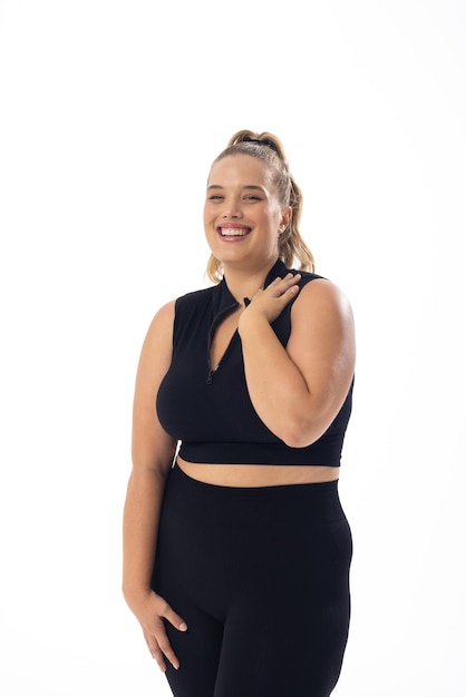 Caucasian young plussize woman in black sportswear stands smiling on white background