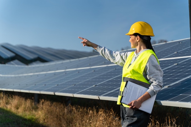 Caucasian young pretty woman professional engineer specialist\
wearing head protection safety outfit pointing away while working\
near solar panels workers concept