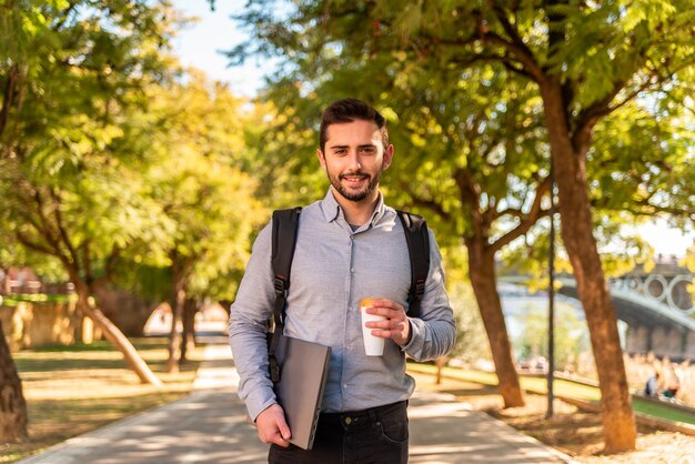 晴れた日に持ち帰り用のコーヒーを飲み、美しい公園で彼のラップトップを運ぶ白人の若い学生と労働者の男。
