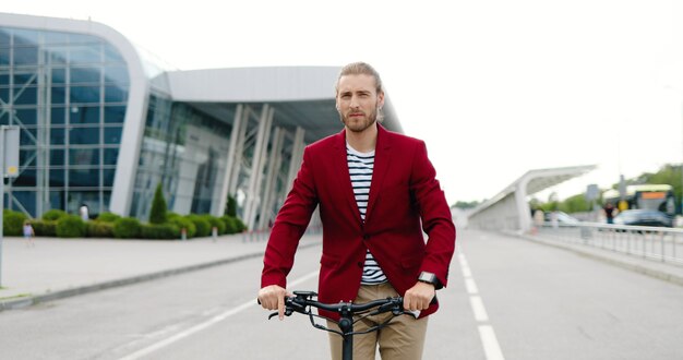 Caucasian young stylish man in red jacket riding electric scooter and looking for time at hand watch. Outdoor. Handsome guy driving and rushing in city. Being late concept.