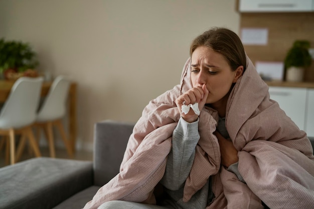 白人の若い女性が咳をして自宅の毛布の下に座っている