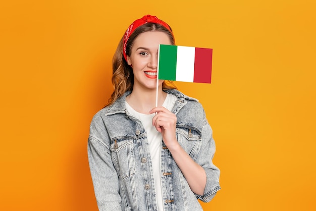 Caucasian young woman in denim jacket covers half of her face with a small italian flag and smiles isolated over orange background student holding italian flag