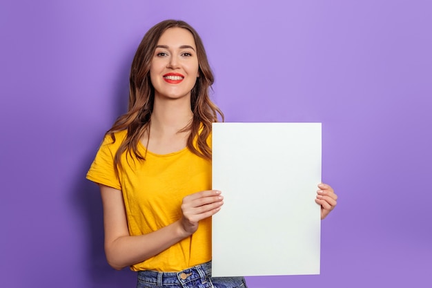 スタジオで薄紫色の背景の上に分離された黄色のTシャツを着て笑顔と空白のポスターを保持している白人の若い女性。デザインのモックアップ