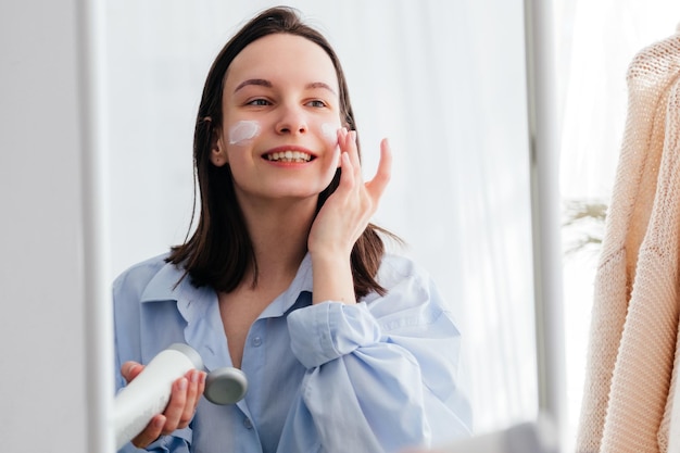 Photo caucasian young woman touching face and applying cream looking in mirror indoors home beauty skincare concept