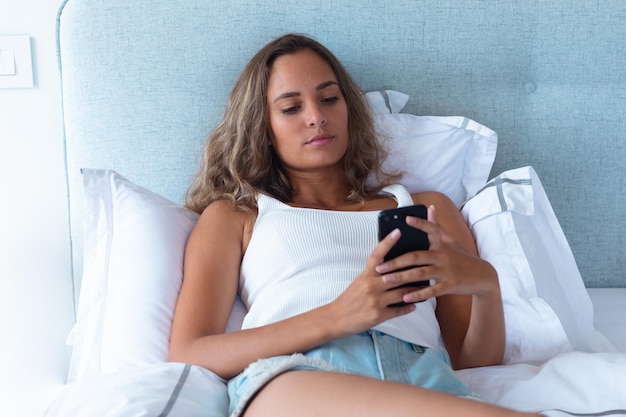 Caucasian young woman using smartphone lying on the bed at home. self-care and relaxation concept