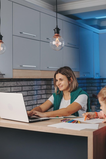 Caucasian young woman working online on laptop with daughter doing her homework in the kitchen inter...