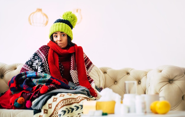 Caught cold. cute young ill woman is feeling cold and sick\
sitting on a couch and being warmly dressed in a sweater, scarf and\
green knitted hat.