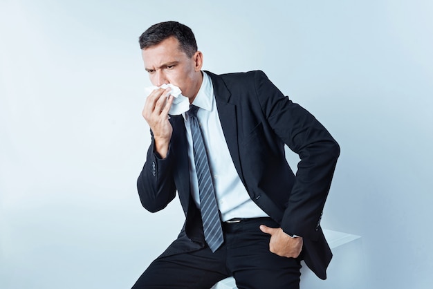 Caught an illness. Chopped shot of a bit confused man sitting on a studio cube and using a paper napkin while wanting to sneeze hard.