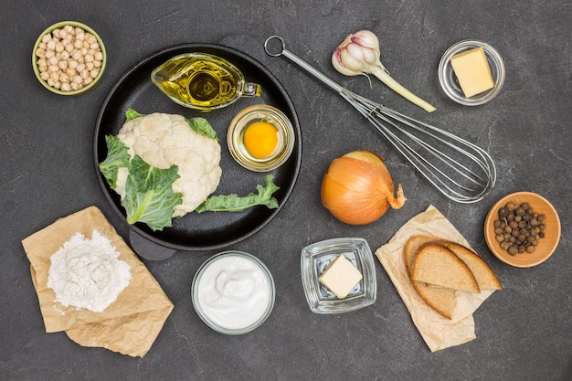 Cauliflower, broken egg and olive oil in frying pan. Whisk, butter, garlic, chickpeas and bread on table. Black background.  Flat lay