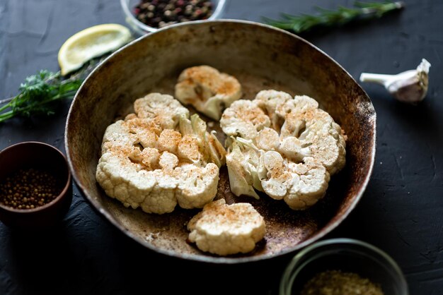 Cauliflower steak cooking Raw cauliflower sprinkled with spices lies in a frying pan Olive oil herbs various spices nearby Dark background