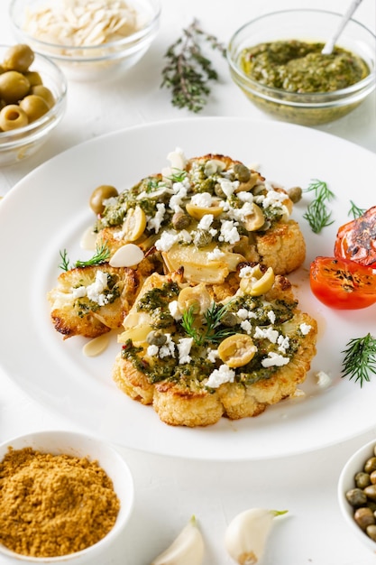 Cauliflower steak with spices chimichurri sauce almond flakes olives fried cherry tomatoes and capers on a white plate Vegetarian food