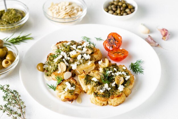 Cauliflower steak with spices chimichurri sauce almond flakes olives fried cherry tomatoes and capers on a white plate Vegetarian food