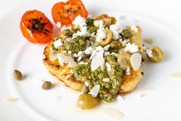 Cauliflower steak with spices chimichurri sauce almond flakes olives fried cherry tomatoes and capers on a white plate Vegetarian food