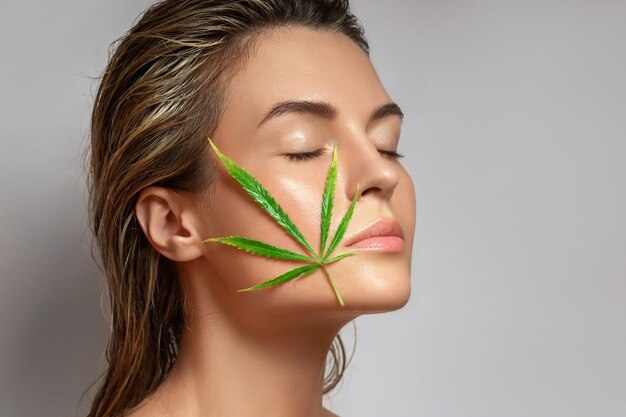 CBD cosmetics concept. Beautiful woman with a cannabis leaf on gray background