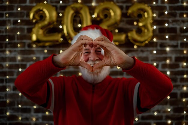 Celebrate event new year for caucasian bearded elderly male making heart shape beautiful senior adult man in Santa hat celebrate holiday at home by night