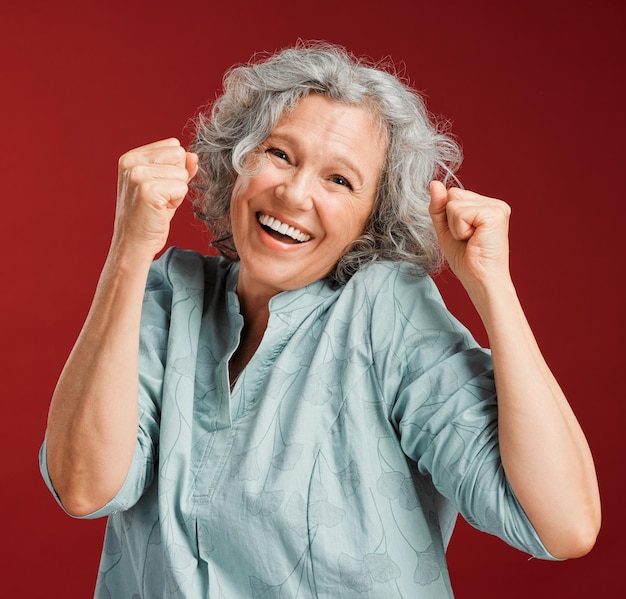 Celebrating cheering and winning with a happy smiling and excited senior female posing in studio against a red background Portrait of a cheerful wow and positive mature female with a fist gesture