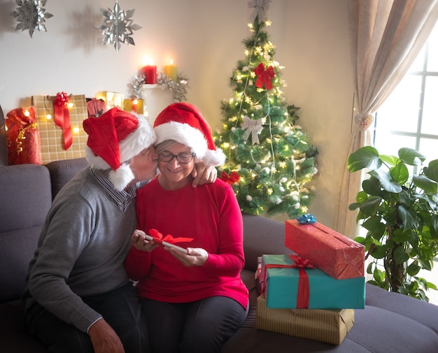 キスと一緒にクリスマスを祝います。贈り物の交換を楽しんでいる美しい老夫婦。彼らはサンタの帽子をかぶっています。背景の美しいクリスマスツリーと家族への贈り物