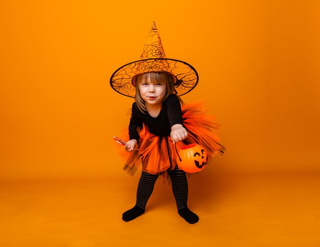 Photo celebrating halloween. little girl dressed as a witch holds a bucket of pumpkins on a yellow background.