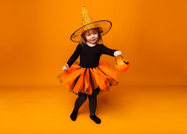 Foto festeggiando halloween. una bambina vestita da strega tiene in mano un secchio di zucche su sfondo giallo.