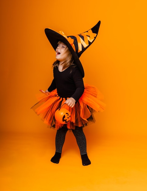 Foto festeggiando halloween. una bambina vestita da strega tiene in mano un secchio di zucche su sfondo giallo.