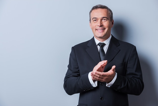 Celebrating success. Confident mature man in formalwear clapping hands and smiling while standing against grey background