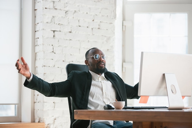 Celebrating win. African-american entrepreneur, businessman working concentrated in office. Looks happy, cheerful, wearing classic suit, jacket. Concept of work, finance, business, success leadership