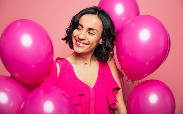 Celebrazione della festa della donna. la giovane donna castana in un vestito rosa sta sorridendo e sta guardando da parte, circondata da palloncini luminosi.