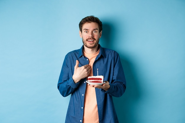 Celebration and holiday concept. Surprised guy holding birthday cake with candle and pointing at himself with disbelief, being congratulated with b-day, blue background.