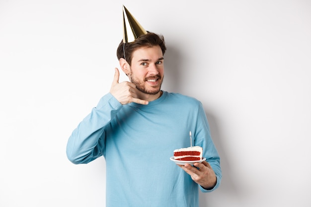 축 하 및 휴일 개념. 파티 모자에 밝은 웃는 남자, bday 케이크와 함께 생일을 축 하, 보여주는 귀, 흰색 배경 근처 전화 제스처 전화.