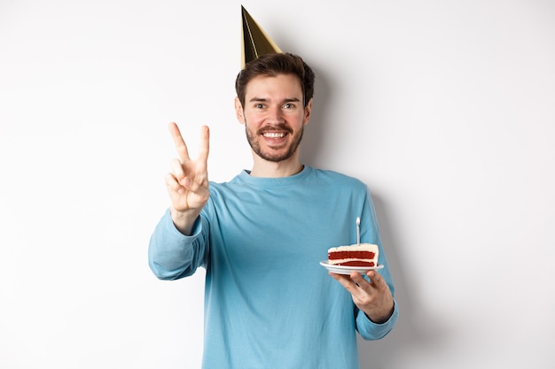 Celebrazione e concetto di vacanze. felice giovane che festeggia il compleanno, scatta una foto con il segno della pace, indossa un cappello da festa e tiene in mano una torta di compleanno, sfondo bianco.