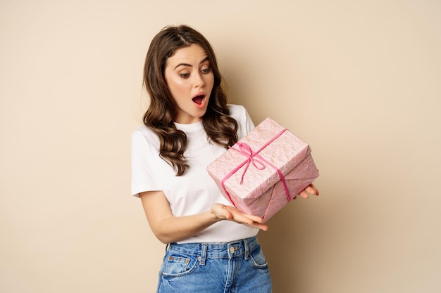 Photo celebration and holidays concept. happy young woman holding gift wrapped in pink box, receive present, looking amazed and surprised, standing over beige background