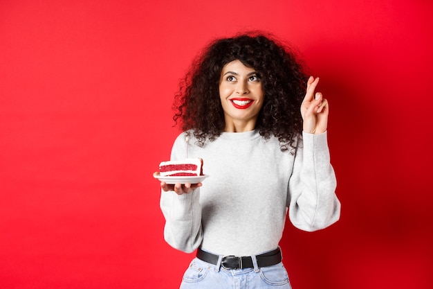 Celebration and holidays concept. hopeful young woman making birthday wish, holding fingers crossed and looking up, holding b-day cake with candle, red wall