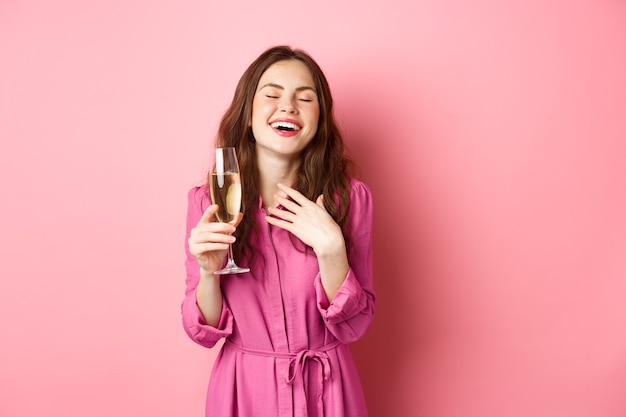 Celebration and holidays concept. Stylish coquettish woman in trendy dress, holding glass of champagne at party, smiling pleased, standing over pink wall.