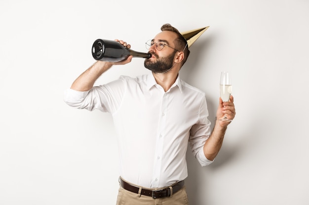 Celebrazione e festività. uomo che beve champagne dalla bottiglia alla festa di compleanno, in piedi su sfondo bianco