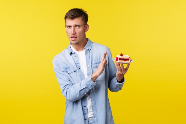 Celebration, holidays and people emotions concept. Reluctant handsome picky guy rejecting dessert, showing stop sign and turn away from cake as dont like sweets, standing yellow background.