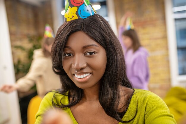 Celebration, joy. Young long haired american girl with toothy smile wearing party hat and friends having fun behind indoors