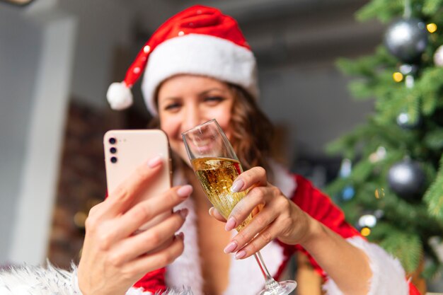 Celebration of new year and Christmas on the distance of the concept. A young woman in a Santa suit and hat is holding a smartphone against the background of a decorated fir tree.