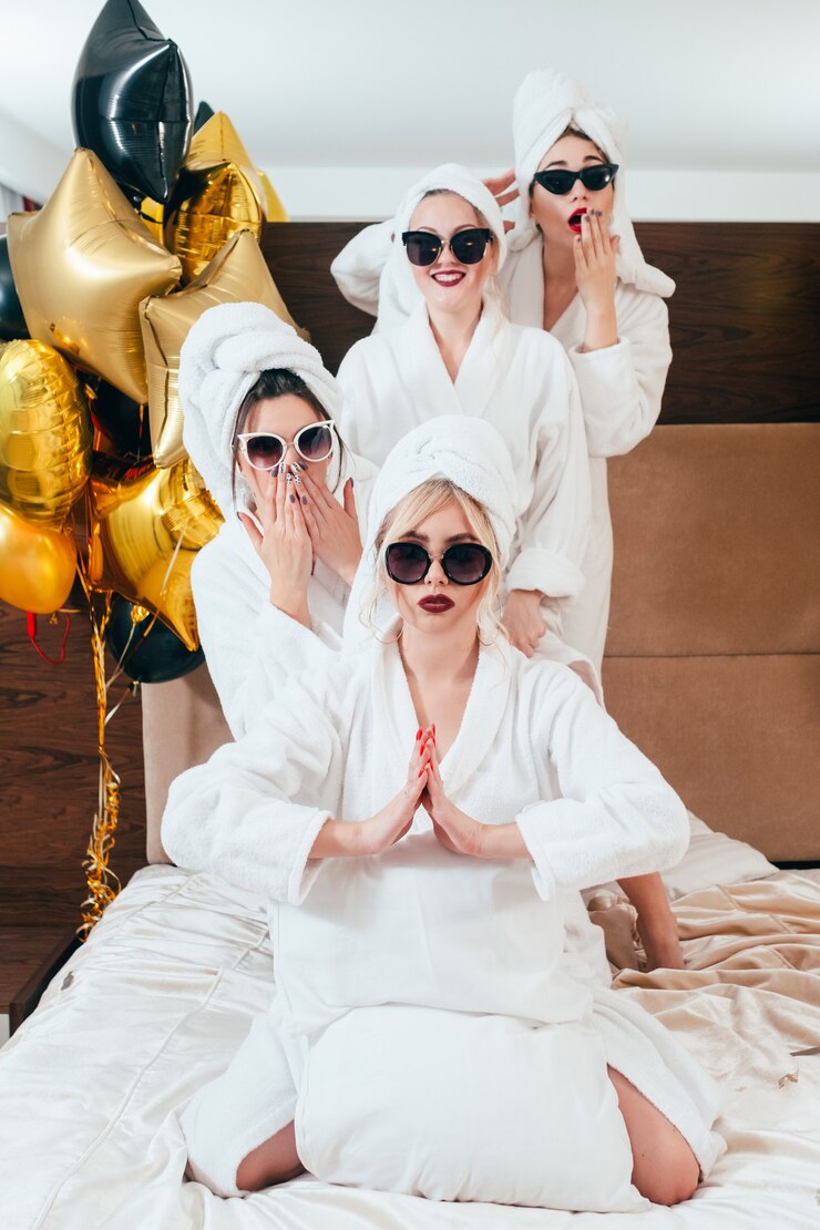 Photo celebration at spa. party fun and relaxation. smiling women posing on bed. sunglasses, bathrobes and towel turbans on.