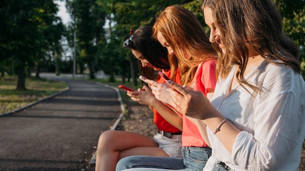 Cell phone addiction group of young multiethnic girls using smartphone sitting in a bench in park