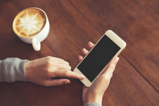 携帯電話のモックアップ画像空白の白い画面。コーヒーショップの机の上で携帯電話を使用してテキストメッセージを持っている女性の手。