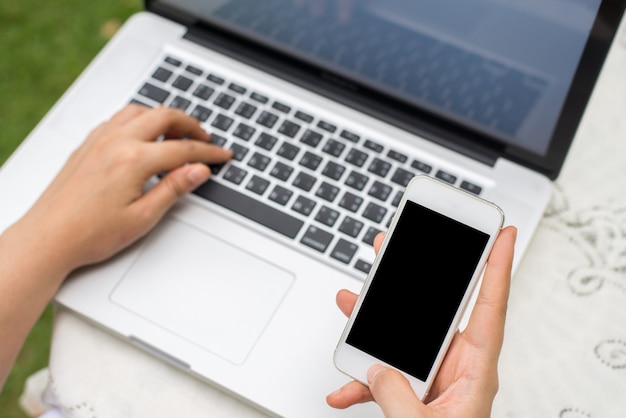 Cell phone with blank screen in woman hands and laptop on wooden table