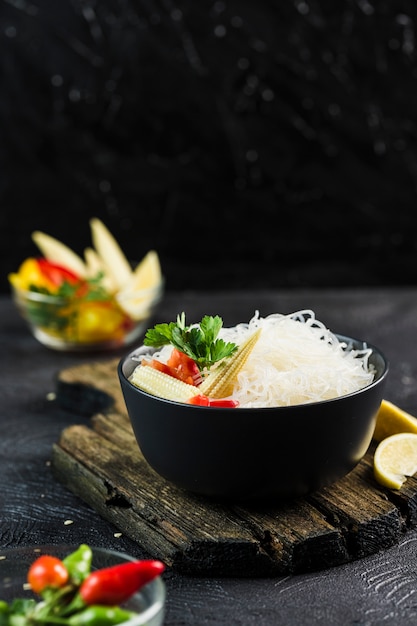 Cellophane rice noodles with vegetables in a black bowl with chopsticks on dark background, side view closeup.